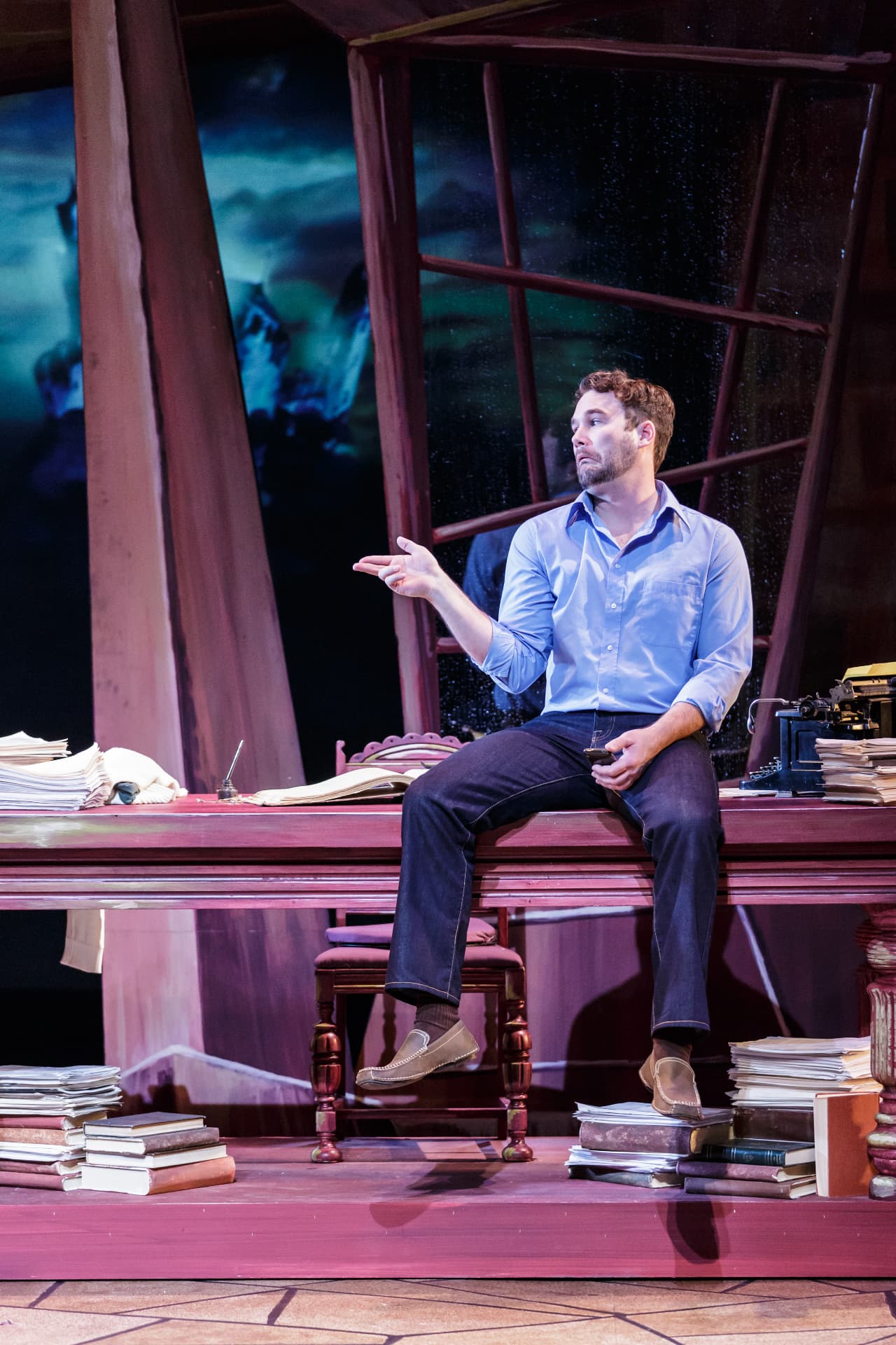 The Understudy — Harry seated on the desk mid-monologue under a rain-streaked window