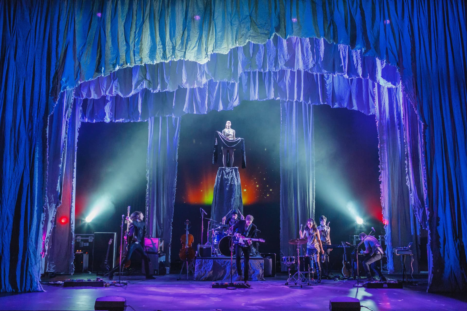 Medea — figure atop a pedestal framed by shredded blue curtains with band below